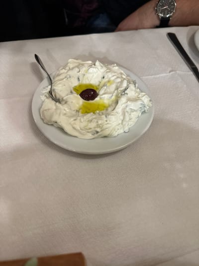 The image displays a close-up, high-angle view of a creamy dip served in a white, round bowl on a dining table, likely in a restaurant in Agios Dimitrios, Greece. The dip, which appears to be a Greek yogurt-based sauce like tzatziki, is white with visible green flecks, suggesting herbs or grated cucumber. A generous drizzle of golden-yellow olive oil pools in the center of the dip, accentuating a single, dark purplish-black olive with its pit. A small, silver spoon rests within the dip, indicating it's ready for consumption.

To the left of the bowl, the lower arm and hand of a person are visible, wearing a sophisticated watch with a dark face, multiple subdials, and a black strap. A silver knife is partially visible at the top center of the frame, resting on the crisp, white, textured tablecloth that covers the entire table surface. A blurred, brown object is partially visible on the far right edge. The lighting suggests an indoor setting. There is no visible text in the image.