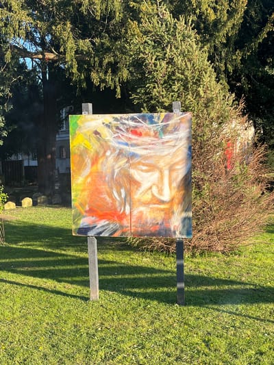 A landscape photograph depicts an abstract painting on a wooden frame, propped up outdoors on a verdant lawn. The painting features swirling brushstrokes of white, yellow, orange, and blue paint, creating a vibrant and chaotic composition. The backdrop consists of lush green foliage, including a large conifer tree on the left and a brown shrub behind the painting. The location is suggested to be in the Borough of Runnymede, United Kingdom, given the prevalence of such landscapes. Sunlight casts distinct shadows of the frame and painting onto the grass, adding depth and contrast to the scene. The composition is primarily focused on the painting and its immediate surroundings.