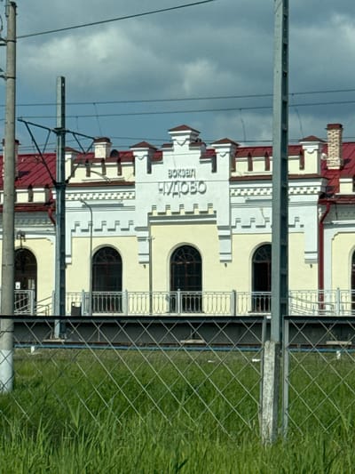 The image shows the Chudovo railway station building in Chudovo, Russia. The station is a two-story building with a light yellow facade and a red roof. The building has a central section with the words "ВОКЗАЛ ЧУДОВО" (Chudovo Station) written in large, white letters. There are arched windows on the ground floor and a decorative railing along the front. In the foreground, there is a chain-link fence and tall green grass. The sky is cloudy. Overhead, there are power lines and a utility pole.