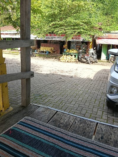 The image displays an outdoor scene in Gembong, Indonesia, observed from a wooden vantage point. In the immediate foreground, a multi-striped mat, primarily blue and green, rests on weathered wooden planks. A vertical wooden beam and a horizontal crossbeam, part of a structural element, are visible on the left, with a yellow-painted edge on the leftmost beam. On the far right foreground, the front bumper, lower grille, and a headlight of a silver-colored vehicle are partially visible.
The midground features a paved surface composed of gray interlocking pavers, with green moss or grass visible in some joints. Scattered shadows are cast across this paved area, indicating direct sunlight.
The background consists of a row of small commercial structures beneath a canopy of lush green trees. A central building displays a red banner or sign, partially obscured, with visible text including "BUTIK" and "PARKIR." Directly in front of this building, a significant quantity of light-colored, spherical fruits, consistent with coconuts or durians, is displayed on a wooden stand and piled on the ground. Several motorcycles are parked to the right of the fruit display. No individuals are visible performing actions or interactions within the scene.