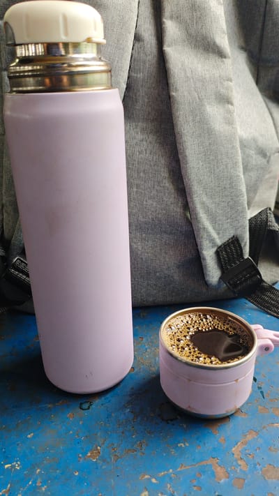 A light purple insulated water bottle and a small light purple mug filled with black coffee sit on a weathered blue surface, likely a table. The coffee mug has a metallic rim and a small handle. Behind the bottle and mug, the corner of a grey fabric backpack is visible, with black straps. The scene appears to be outdoors, given the sunlight and the textured surface, possibly in a park or cafe setting in Goczałków, Poland. There are no people or animals present in the image.