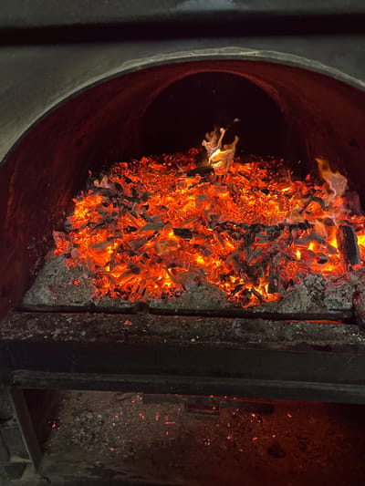 The image displays the interior of an arched combustion chamber, likely an oven or large hearth, located in the city of Dobrodan, Bulgaria. A substantial bed of actively glowing red and orange embers, intermingled with grey ash and dark, partially carbonized wood fragments, occupies the upper two-thirds of the visible space. Several small, yellow-orange flames are discernible, primarily towards the center-right of the ember mass. The internal surfaces of the chamber appear dark and stained. In the lower foreground, a horizontal, dark-colored metallic grate or barrier, encrusted with ash, spans the width of the opening, separating the main ember bed from a lower collection area where sparse ash and faint glowing particles are visible. No individuals are present in the scene.