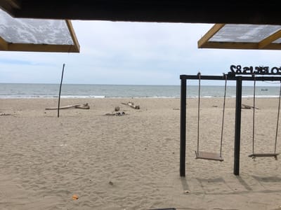 A serene beach scene unfolds under a cloudy sky, possibly indicating an overcast day or late afternoon. The foreground is dominated by sand, marked with the gentle ripples of the tide and scattered with small pieces of debris and a few dry leaves. Two wooden swings, suspended by ropes from a dark, sturdy frame, are the prominent objects. They hang empty, casting faint shadows on the sand.
In the mid-ground, the ocean stretches out, its surface a muted gray-blue, reflecting the subdued light. Small waves gently lap at the sandy shore. A few pieces of driftwood lie scattered on the beach, one notably near a single dark pole stuck vertically in the sand. Further back, a small, dark boat with a few figures can be seen on the water.
Above the beach, a wooden structure is visible, with two transparent, possibly plastic, coverings supported by yellow wooden beams. These coverings are angled, suggesting they are part of a roof or awning, offering shelter from the elements. The overall impression is one of tranquility and a peaceful, albeit somewhat unpopulated, coastal setting. Visible text on the swing frame reads "SO BRAS83" in stylized black lettering. The location is Keudekruenggeukueh, Indonesia.
