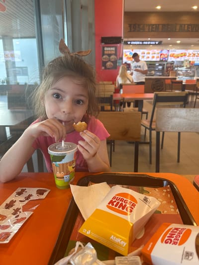 A child is seated at an orange table at a Burger King restaurant in Manisa, Turkey. The child has light brown hair with a bow and is wearing a pink shirt. They are holding a chicken nugget in their left hand and drinking from a decorated cup with a straw in their right hand. On the table is a tray with Burger King food containers, including a box of nuggets, a box of fries, and a cup. Sticker sheets featuring cartoon animals are also on the table. In the background, other tables and chairs are visible, along with a Burger King ordering area. The restaurant's slogan "1954 TEN BERIJALEVD" is displayed on the wall.