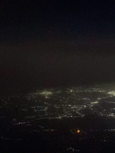A dim, dark, and slightly grainy image reveals a blurred scene. The left side appears predominantly dark, with subtle variations in shading. Moving right, there's a shift as light elements appear. These are mostly vertical and irregular in shape, suggesting possible streaks or reflective surfaces, perhaps raindrops on glass at night with a single light source visible in the dark beyond.