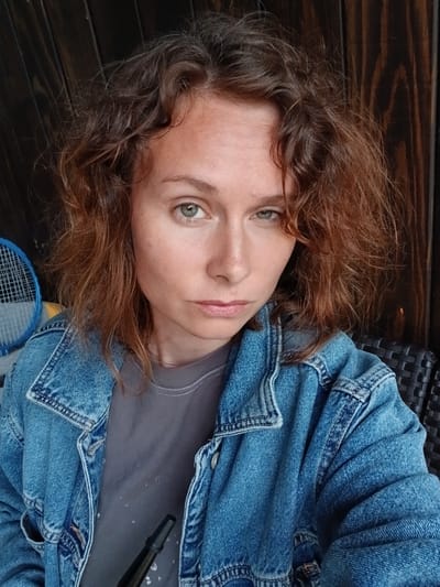 In Verkhneye Dubrovo, Russia, a medium shot captures a woman with auburn, curly hair and green eyes. She is wearing a denim jacket over a grey t-shirt. Her expression is neutral. The background consists of wooden planks and a wicker chair. To her left, a portion of a blue and white tennis racket is visible. A black object, possibly a hookah mouthpiece, is held in her lower right quadrant.