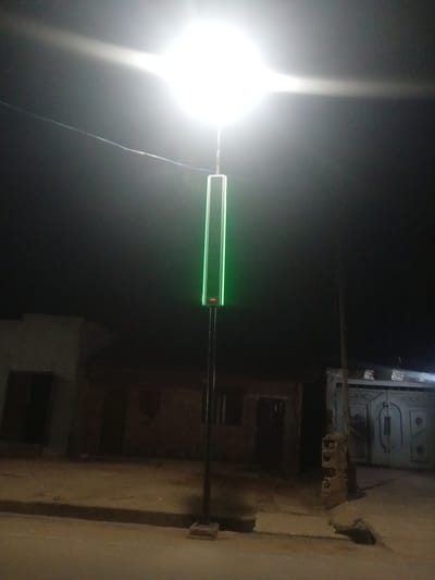 In the city of Tundun Wada West, Nigeria, a tall, slender pole stands centrally in the foreground, topped by a bright, luminous light that washes out the upper portion of the image. Mounted on the pole, below the primary light source, is a vertical, rectangular panel emitting a vibrant green light. The panel has subtle red markings at its base. The surrounding environment is dimly lit, with the glow of the light illuminating a dusty, uneven ground. To the left and right of the central pole are buildings constructed with brick. The building on the left has a dark opening that appears to be a doorway. The building on the right features a large, ornate metal gate with intricate patterns. The overall impression is a street scene at night, illuminated by artificial light.