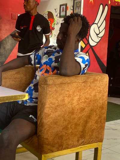 A medium shot captures two Black African men indoors against a brightly colored and patterned background. The man closer to the camera is seated, wearing a blue and white patterned shirt and black shorts with "Play" printed on them. He is leaning into a brown cushion attached to a yellow chair, his hand raised near his head as if in thought or discomfort. The second man stands to the left, wearing a black shirt with "24/7" printed on it and a red cap. He holds a phone. The backdrop includes a red wall with black lines, a white peace sign, and framed pictures. A mural featuring mushrooms and an orange-filled brown bowl, likely food-related, is also visible on the white wall. Artificial green turf is present near the top right of the frame. The image appears to be taken at eye level, with the men as the central subjects. There are no other discernible markers to conclude it is Tukur Tukur, Nigeria.