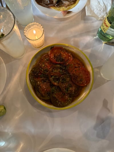 The image displays a dinner table setting at a restaurant in Guadalajara, Mexico, seen from an overhead perspective under low light conditions. Central to the frame is a white oval bowl with a bright yellow rim, containing numerous slices of red and dark green heirloom tomatoes, likely roasted or marinated, resting in a dark liquid. These tomatoes are garnished with chopped green herbs and white specks, possibly salt or cheese.

To the left, two clear glass tumblers hold light-colored beverages with ice, one showing a visible lime wedge. A small, lit white candle in a fluted glass holder casts a warm glow between these glasses and the tomato dish.

In the upper right quadrant, a white plate partially shows a taco-like dish featuring meat on a tortilla. Next to this plate is a crumpled white paper napkin. A green glass bottle of "Topo Chico" mineral water is also visible in the upper right. A small, fresh lime wedge rests directly on the white tablecloth in the lower left corner. The table is covered with a white, slightly wrinkled tablecloth, reflecting light from the candle and other unseen sources. No individuals are visible.
