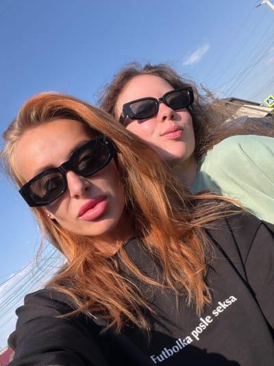 This image depicts two young adult females taking a selfie outdoors in Yemelyanovo, Russia, on a bright, sunny day with a clear blue sky. The woman in the foreground, with long reddish-orange hair, full pink lips, and a tanned complexion, wears black rectangular sunglasses and a black long-sleeved top that bears the white Latin script "Futbolka posle seksa." Her left ear features a small stud earring. Positioned behind and to her right, a second woman with wavy brown hair wears similar black rectangular sunglasses and a light green crew-neck sweatshirt, with her lips slightly pursed. The background reveals a suburban environment with numerous overhead power lines, a utility pole, the partial view of residential building roofs (one dark, one reddish), and a yellow diamond-shaped pedestrian crossing sign. Strong sunlight creates distinct shadows.