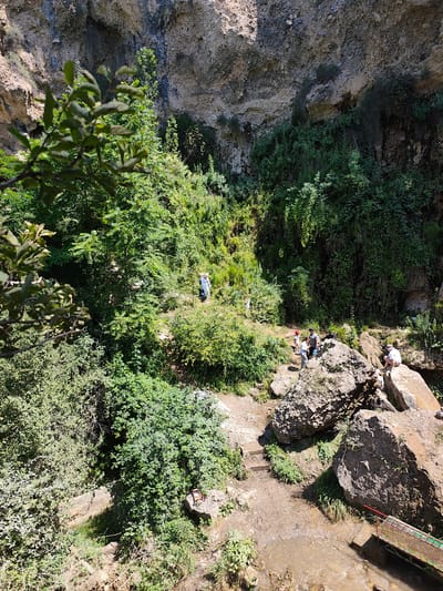 In Arstanbap, Kyrgyzstan, a natural landscape unfolds with a prominent cliff face dominating the background, dotted with vegetation. In the mid-ground, several individuals are scattered across a rocky and earthy terrain, surrounded by lush green foliage. One person in a blue shirt and white pants stands near the center, while a small group of three people, one in a red cap, are gathered on a large boulder to the left of the center. Further to the right, another individual is seated on a rock. The foreground features a muddy stream flowing beneath a small, vibrantly colored bridge with red, yellow, and green railings. The overall scene depicts a natural, outdoor setting with people enjoying the environment.