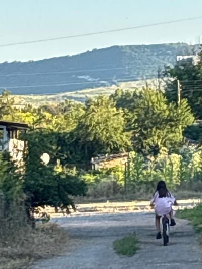 The image depicts a young person riding a bicycle on a dirt road in a rural setting. The person is viewed from behind and appears to be wearing a light-colored shirt. The environment is characterized by lush green trees and vegetation, with rolling hills in the background under a clear sky. A simple building structure is visible on the left side of the frame. The scene suggests a peaceful, outdoor activity in a natural landscape. The image is taken in Enevo, Bulgaria.
