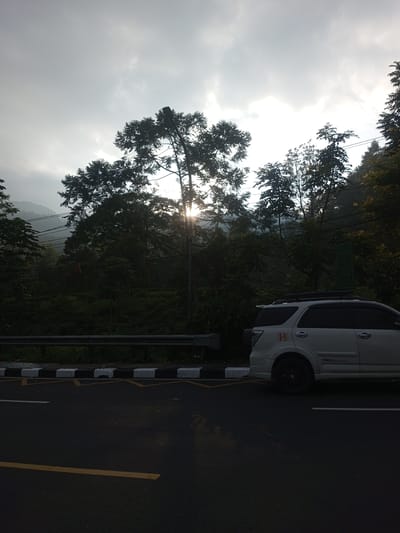The image depicts a street scene near Taunggyi, Myanmar. A white SUV with a roof rack is parked on the right side of the road, near a black and white striped curb. A metal guardrail runs along the edge of the road, separating it from a forested area. The trees obscure the view of a mountain in the background. Sunlight shines through the trees. The sky is overcast. The road has a yellow line.