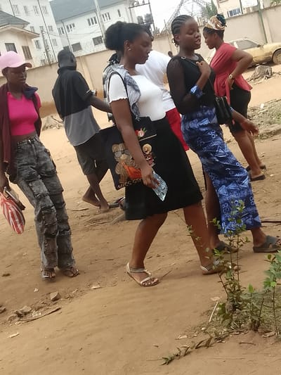 The image depicts an outdoor scene in Umuokpu, Nigeria, focusing on a group of five individuals walking on an unpaved dirt path. In the foreground, from left to right: a female wearing a pink baseball cap, a pink crop top, a dark-colored cardigan, and distressed grey jeans, carrying a red and white striped bag; a male in a black and grey t-shirt and grey shorts, walking barefoot; a female wearing a white ruffled-sleeve top, a black skirt, a light grey patterned scarf over her shoulder, carrying a black tote bag with a bear design, and holding a clear plastic bottle; a female wearing a black long-sleeved top and a blue and white patterned ankle-length skirt, carrying a small black handbag; and a partially visible female in a red top, black skirt, and a patterned headwrap, wearing a watch on her left wrist. All individuals appear to be walking towards the right side of the frame. The ground is brown dirt with small rocks and minimal vegetation, including a small green plant in the lower right foreground. In the background, modern-style, light-colored multi-story residential buildings with dark roofs are visible behind a fence topped with barbed wire. A light-yellow, older model car is parked or abandoned further back to the right, near a tree stump. The sky is not visible.