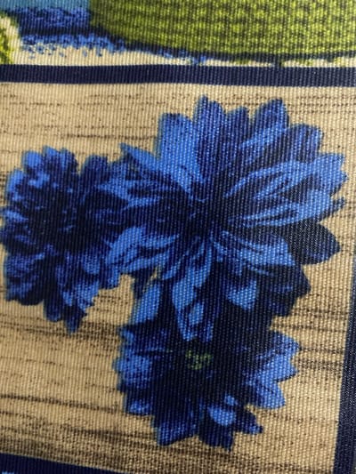 This is a close-up, overhead shot of a fabric with a floral pattern. The pattern features large, stylized blue flowers with layered petals. The flowers are set against a textured, beige background with vertical lines. To the left of the flowers, there is a vertical stripe of dark blue and a patch of green, possibly representing leaves or another floral element. The fabric appears to be woven, with visible threads creating a textured surface. The lighting is even, and there are no prominent shadows, suggesting an indoor or overcast setting. The image is focused on the detail and texture of the fabric, emphasizing the vibrant blue of the flowers. There is no discernible activity or event taking place, as it is a still image of a textile. No text is visible in the image. The context provided, Mitoc, Moldova, suggests this fabric might be part of clothing, home decor, or a craft item from that region.