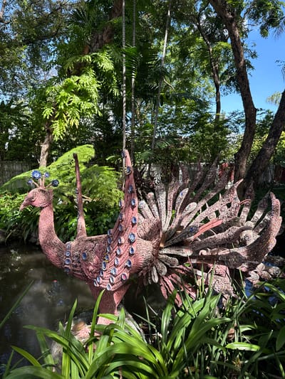 A mosaic-covered peacock sculpture stands within a garden setting in Samrong Nuea Subdistrict Municipality, Thailand. The sculpture is pinkish-red, adorned with blue and white circular mosaics that resemble eyes along its neck and tail feathers. The peacock is positioned near water, potentially a pond or small stream, surrounded by lush green vegetation, including various plants and trees. The scene suggests a garden or park setting, likely a tourist spot. The photo is shot with a wide-angle lens under natural sunlight, casting shadows and highlighting the textures of the mosaic and the foliage.