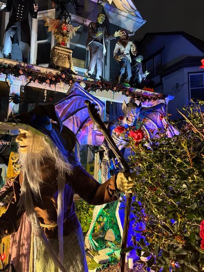 The image displays a residential house in Weehawken, United States, heavily decorated with Halloween-themed props at night. The scene is illuminated by various artificial colored lights including blue, purple, green, and red.
In the foreground, a life-sized witch figure with a long white beard, brown robe, and a pointed hat holds a gnarled staff in its right hand. To its right, a large winged bat-like creature figure with glowing eyes, sharp teeth, and spread wings is visible, adorned with red roses and bathed in purple and blue light. A dense green bush with several red roses occupies the immediate right foreground, partially obscuring the bat creature. The ground level exhibits artificial green lighting and scattered white decorative objects.
On the house's porch or balcony level, a series of static figures are arranged. From left to right, these include a figure in a dark formal coat, a gargoyle sculpture on a pedestal with glowing red eyes, a green-skinned Frankenstein's monster figure, a fur-covered werewolf figure with outstretched arms, and a smaller green-skinned figure. A garland of dark flowers and string lights is draped along the roofline above these displays.
The background shows the upper facade of the decorated house with windows, and an adjacent structure with a pitched roof, partially illuminated. The sky is dark, indicating nighttime.