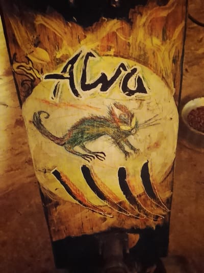 This image depicts a wooden object, identified as a skateboard or longboard deck, presented at an angle, within the city of Bernal Este, Argentina. The deck features intricate hand-painted artwork. Central to the design is a stylized, feral-looking creature resembling a cat, rendered with green, brown, and red fur-like texture, prominent whiskers, and intense yellow eyes, depicted in a low, forward-moving posture. Above this creature, the word "Awa" is painted in thick, black, irregularly shaped letters, outlined in a lighter shade. Both the creature and the text are enclosed within a large, irregular light yellow oval. Surrounding this oval, dark yellow and orange brushstrokes, suggestive of flames or abstract patterns, extend onto the exposed wood grain of the deck. Below the creature, within the yellow oval, several dark, vertical, rib-like or claw-mark elements are visible. The underlying material is natural wood, showcasing its grain and texture in the unpainted areas. The scene is illuminated by warm, indirect light, indicating an indoor or sheltered setting. In the blurred upper-right background, a small, white ceramic bowl containing reddish-brown granular material rests on a light brown surface. Dark hardware, consistent with a skateboard truck assembly, is partially visible at the bottom edge of the deck. No individuals are present.