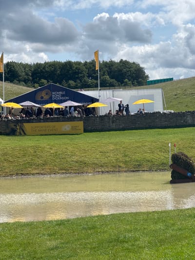 The image shows a view of a large grassy area with a water obstacle in the foreground, and a large tent in the background. The water obstacle is a small pond with a wooden frame and brush surrounding it. There are two red flags marking the obstacle. The tent is blue with yellow signage that reads "WOMBLE BOND DICKINSON" and "SUPPORTERS OF THE MAIN EVENT." In the background, there are several people gathered near the tent, and a line of green trees can be seen in the distance. The sky is mostly blue with white clouds. The image was taken at the Bramham International Horse Trials, located near the village of Bramham, West Yorkshire, United Kingdom.