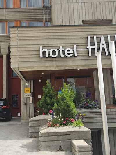 The image shows the exterior of the Hotel HAUS post-fire in St. Moritz, Switzerland, with a sign above the entrance reading "hotel HAUS". The hotel's facade is made of concrete with horizontal grooved detailing. To the left of the entrance, a dark-colored car is parked, and the number "1" is visible on a wall. Adjacent to the entrance, a yellow sign indicates the hotel is a "SMARTY HOTELS MEMBER". A small green Christmas tree is planted in front of the entrance, surrounded by various flowering plants. The entrance itself features a wooden door and is under a covered walkway. The weather appears to be fair.