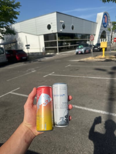 A person's hand holds two energy drink cans in front of a blurred modern building with a large blue circular logo. The can on the left is red and yellow with a white bull graphic and text in French. The can on the right is white and silver with text in French, including "WHITE EDITION" and "ENERGY DRINK". In the background, a paved parking lot is visible with white parking lines. Several cars are parked or driving in the distance. The building appears to be a retail establishment with large windows and a distinctive arched entrance. The sky is clear and blue. This scene is located in Alès, France, likely in front of a Leclerc hypermarket, given the logo.