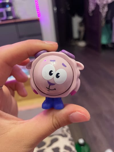 A close-up shot shows a person's hand holding a small, pink, cartoonish toy sheep. The toy has large white eyes with black pupils, a simple smiling mouth, and purple accents on its ears and head. It is wearing blue pants. The background is blurred, showing a dark room with some purple lighting. The image does not contain enough information to determine the location in the city of Borisova Griva, Russia.
