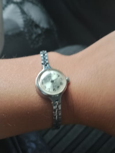 A close-up shot shows a wrist wearing a vintage silver-colored analog watch. The watch has a circular face with a light-colored dial and dark hands. The band is made of interlocking silver links. The wrist is bare, and the skin appears light-toned. The background is blurred and dark, suggesting an indoor setting, possibly a car interior or a dimly lit room. The image is a personal photograph taken with a mobile phone, likely in a casual setting, not in a specific city location.