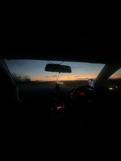 The image captures a view from inside a vehicle, looking out through the windshield during sunset or twilight. The scene is primarily dark, with the car's interior visible mostly in silhouette and illuminated by internal lights.

The sky, seen through the windshield, dominates the exterior view. It displays a gradient of warm hues, transitioning from soft orange and yellow near the horizon to cooler blues and grays higher up, indicative of a sunset or sunrise. Some clouds are visible against the colorful sky. Below the horizon, the landscape appears dark and indistinct, possibly a road or open terrain, with scattered distant lights suggesting other vehicles or distant structures.

Inside the car, a rearview mirror hangs from the top center of the windshield. To its right, a smartphone or navigation device is mounted, showing a map interface with a blue route line. On the right side of the frame, parts of the car's dashboard are visible, including a steering wheel and red illuminated gauges behind it. Further to the right and lower, illuminated controls on the center console or dashboard can be seen, with "A/C OFF" clearly legible in red text.

The image suggests the car is in motion or stopped on a road, with the driver's perspective implied. The time of day is evening, specifically sunset or twilight. The location context is Pucklechurch, United Kingdom, which aligns with the rural or semi-rural landscape visible. No specific individuals are discernible in the photograph.