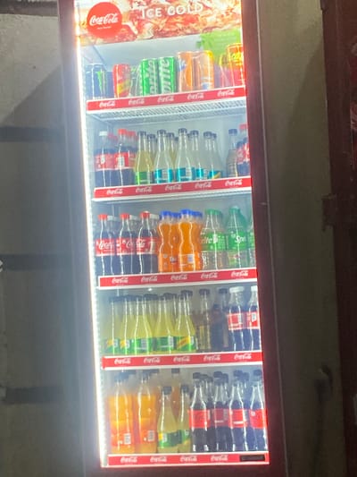 A refrigerated display case stocked with various Coca-Cola products and other soft drinks is visible. The Coca-Cola logo is prominently featured on the cooler and product labels. The bottles and cans are arranged in rows, indicating a commercial setting. There is no evidence or indication to determine the location or suggest it is in Tukur Tukur, Nigeria. The image shows beverages in a cooler.