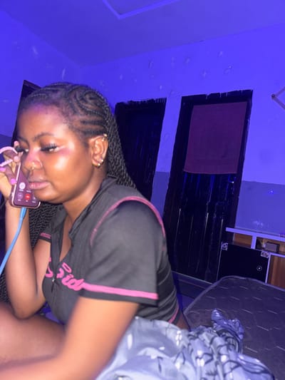 An individual is depicted indoors in Gbongan, Nigeria, seated and holding a smartphone to her left ear, engaged in a phone call. The subject is a female with dark skin, braided hair, and multiple ear and nose piercings. She wears a black, short-sleeved top with pink trim and partially visible lettering. A blue wired earphone cable extends from the phone. The indoor environment is illuminated by a strong blue ambient light. In the background, two dark doors are visible, with a reddish-brown towel draped over the door on the right. The walls are light-colored with visible surface texture. A patterned blanket partially covers her lower body, which rests on a dark mattress or bed base. A white shelf with a dark electronic device is partially visible on the far right.