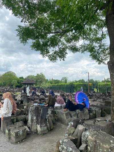 The image shows a group of people sitting on and around large gray stone blocks, which are likely part of a historical ruin or temple complex. The stone blocks are covered in moss and lichen, indicating age and exposure to the elements. The scene is set against a background of lush green vegetation and a cloudy sky. A large tree with green leaves casts shade over the people. Some people are wearing head scarves and traditional clothing. A few people are standing in the background, and one person is lying on a stone block with a large purple umbrella over their head, possibly taking a nap or enjoying the shade. The scene is located in Kecamatan Prambanan, Indonesia, which is known for its ancient Hindu temples. The coordinates -7.75 latitude and 110.492 longitude place the scene within the vicinity of the Prambanan temple complex, suggesting that the image was taken within or near the temple grounds.