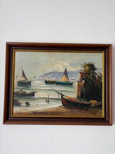 The image is a painting of a coastal scene with several sailboats on the water. In the foreground, a red and green boat is pulled up on a sandy beach. To the right of the boat is a small stone building with a dark, arched doorway and trees growing around it. In the middle ground, there are more sailboats on the water, some with their sails up. In the background, there are hills and a cloudy sky. The painting is framed in a dark brown wooden frame with a gold inner border. The signature "G. Parath" is visible in the lower left corner of the painting. The painting is hanging on a white textured wall. There is no identifiable location in Osnabrück, Germany depicted in this painting.