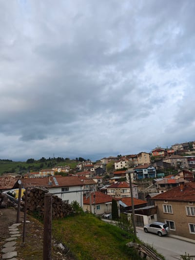 The image shows a view of the city of Kasak, Bulgaria. The scene is taken from an elevated position, likely a hillside or a building. The houses are built on a slope, with rooftops of various colors, predominantly red tile, visible. There are multiple houses, some with brick exteriors, and various colorations of their walls. A single silver car is parked on a road. The sky is filled with grey, overcast clouds. There is a green, grassy area in the foreground, and a pile of logs is stacked on the left. There is a metal support structure in the foreground. A hillside covered in green vegetation and scattered trees can be seen in the distance.