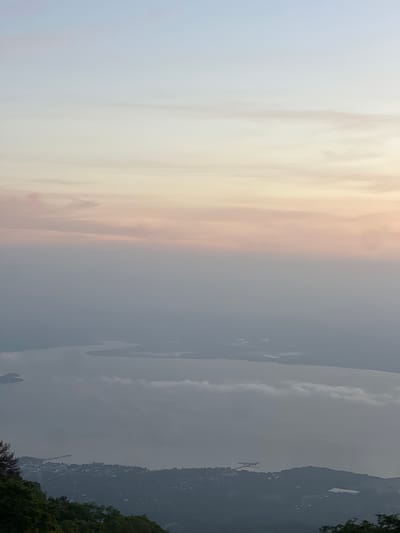 This is a high-angle, vertical, full-frame shot of a hazy, diffused dawn sky over a body of water and a coastal town. The sky transitions from a pale, almost white blue at the top to soft pinks and oranges as it approaches the horizon. A layer of mist or low clouds hangs between the sky and the landscape, obscuring much of the detail. Below the mist, a wide, calm body of water, likely a bay or large lake, is visible. Islands or peninsulas break the surface of the water, their shapes softened by the haze. Along the shoreline, a dense cluster of buildings, indicative of a town or city, is spread across the land. Roads and structures are visible, though indistinct due to the distance and atmospheric conditions. The foreground features the dark, shadowed canopy of lush green trees, suggesting the viewpoint is from a higher elevation, possibly a mountain or hill overlooking the scene. The location is identified as El Cacao, El Salvador.