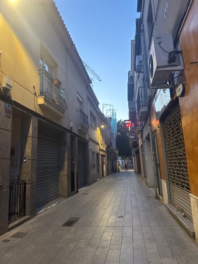 The image depicts a narrow street in Sant Boi de Llobregat, Spain, possibly in the evening given the lighting conditions. The street is paved with rectangular tiles. Buildings line both sides of the street, with the one on the left being a lighter color and featuring balconies and closed roll-up doors on the ground floor. The building on the right is a darker orange color and also has closed shutters.
In the distance, a lone figure, appearing to be a person wearing a red top, walks away from the camera down the street. Above the buildings, a digital clock displays "6 07" with the word "Sancho" above it. A construction crane is visible further in the background. Air conditioning units are mounted on the exterior walls of some buildings. The overall atmosphere is quiet and somewhat commercial, with the closed storefronts suggesting the end of business hours.