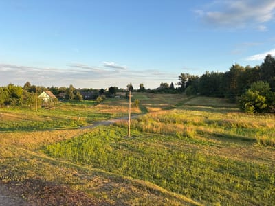 The image depicts a rural landscape with several houses and trees under a clear blue sky with scattered clouds. The scene appears to be a village setting within Краснабярэжскі сельскі Савет, Belarus. In the foreground, there is a path winding through a grassy field. To the left, a light green house with a dark roof is visible, surrounded by trees and a fence. Further in the distance, more houses and dense greenery can be observed. A wooden utility pole stands in the middle of the field, with power lines extending across the landscape. The lighting suggests it is either early morning or late afternoon, with the sun casting long shadows across the field, highlighting the textures of the grass and vegetation. The overall atmosphere is tranquil and serene, characteristic of a rural Belarusian village.