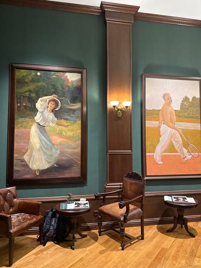 The image depicts an interior scene, likely a gallery or a well-appointed room. Two large framed paintings are the focal point. The painting on the left shows a woman in a white dress playing tennis in an outdoor setting with trees and a body of water. The painting on the right features a shirtless man in white pants holding a tennis racket, walking on a tennis court. Between the paintings is a wooden pillar with a decorative light fixture. In the foreground, two leather armchairs are positioned next to small round wooden tables. One table holds a glass of water, a cup of coffee, and some magazines, with a black backpack placed on the floor nearby. The other table also displays magazines. The walls are painted a deep green, and the floor is polished hardwood. The overall impression is one of elegance and a focus on the sport of tennis. The image is not located in the city of Zhukovka, Russia.
