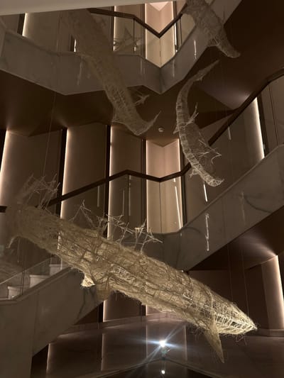 Four sculptures of fish hang in front of a beige and dark-brown geometric ceiling, with light shining in the upper-right corner. The fish are constructed of a pale, fibrous material with an open, cage-like structure. The location may be El Gouna, Egypt.