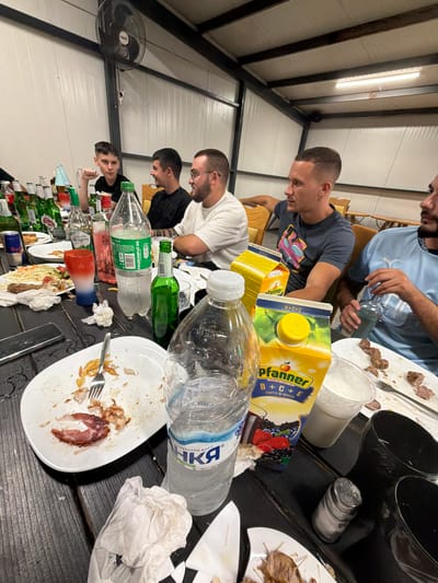 A group of young men are seated around a dark wooden table in what appears to be an indoor, casual dining area. The environment suggests a gathering, possibly a party or meal, given the presence of food remnants, bottles, and drinks on the table. The lighting is somewhat dim, with overhead fluorescent lights visible. In the background, a large industrial-style fan is mounted on the ceiling. The men are casually dressed, and some appear to be engaged in conversation. On the table, there are plates with leftover food, several bottles of alcohol and soft drinks, juice boxes, and glasses. Some of the bottles are empty or partially consumed. There is also a salt shaker and crumpled napkins. The overall scene depicts a post-meal environment, suggesting a relaxed and informal social event. The location is specified as Chavei, Bulgaria.