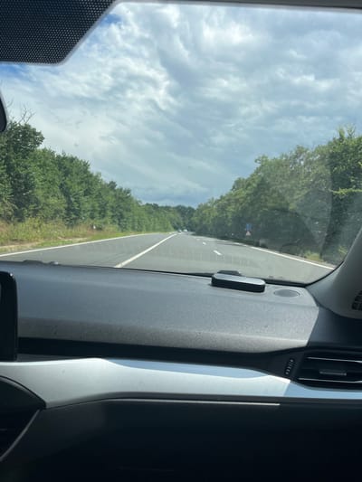 The image shows a view from inside a car traveling on a road. The road is paved with white lane markings and is bordered by dense green trees on both sides. The sky is cloudy, with patches of blue visible. The interior of the car is visible in the foreground, with the dashboard and part of the windshield. There is a small black electronic device on the dashboard. The location is specified as Cotu Morii, Romania.