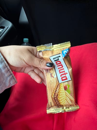 A close-up shot shows a person's hand holding a "Hanuta" chocolate wafer bar. The bar is wrapped in gold foil and features a prominent red and white label with the brand name. The hand has black nail polish and a light blue ring on one finger. The background is a dark, out-of-focus interior, likely a car, with a glimpse of a water bottle with a blue cap. The hand and the wafer bar are positioned over a bright red fabric surface. The image does not contain any discernible elements that would allow for identification of the location as Gornji Vakuf-Uskoplje, Bosnia and Herzegovina.