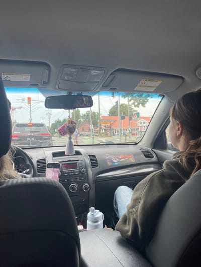 The image shows the interior of a vehicle from the rear passenger seat, looking forward through the windshield at an urban intersection in Saanich, British Columbia, Canada. The vehicle is stationary, facing north at the intersection of Shelbourne Street and Feltham Road, as indicated by visible landmarks and a red traffic signal.
In the foreground, the driver's head, displaying light-colored hair, and their left shoulder are visible. A front passenger is seated to the right, observed from behind, wearing a dark olive green hoodie and light blue jeans; their brown hair is tied back. The vehicle's dashboard features a central infotainment system. A smartphone is mounted to the left of the system, displaying an interface with pink and purple tones. A light blue and white water bottle is placed in a central cupholder. A multi-colored sticker depicting a mountainous landscape is affixed to the passenger side dashboard. The rearview mirror is adorned with two pink fuzzy dice, a heart-shaped charm, and a white tassel, and includes a compass displaying 'N' for North. The windshield exhibits moisture, suggesting recent or ongoing precipitation.
Through the windshield, a dark-colored SUV, identifiable by a "Jeep" logo on its rear, is stopped directly ahead in the same lane. To the right, commercial buildings feature red-tiled roofs and light-colored facades. Signage for "LOADED PLUS CHICKEN & BURGERS" and "KFC" is clearly visible on these buildings. Multiple pedestrians are visible on the far side of the intersection. A large construction crane is present in the distant left background, and the sky is overcast.