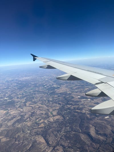 From an airplane window, a wing extends over a landscape of varied terrain. The sky transitions from deep blue to a lighter haze on the horizon. Below, the ground displays a mix of textures, possibly including fields, forests, and rocky areas. A winding river cuts through the landscape, adding to the visual complexity of the view from above.