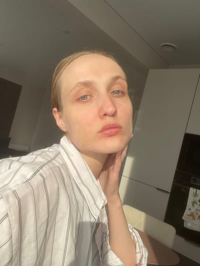 The image presents a selfie of a light-skinned individual with blonde hair and blue eyes. The person is wearing a white striped shirt and is positioned at an angle, with their right hand touching their neck. The setting appears to be indoors, likely a kitchen, given the presence of cabinets and a kitchen towel visible in the background. The lighting suggests daytime. There is no information to confirm that this image was taken in Sepych, Russia.