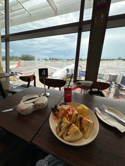 The image depicts a table in an airport lounge at Phuket International Airport, Mai Khao, Thailand. On the dark wooden table, there is a plate of food with a sandwich, salad, and potato chips, a can of Coca-Cola with a straw, and a pair of headphones. In the background, through a large window overlooking the tarmac, several airplanes are visible, along with airport infrastructure like jet bridges and service vehicles. The sky is overcast, suggesting it might be a cloudy day. A table number '4' is also prominently displayed on the table.