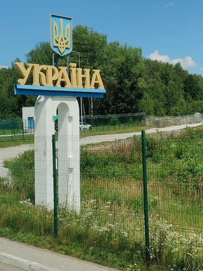 The image shows a border crossing sign with the Ukrainian emblem and the word "UKRAINE" in large, yellow, 3D letters. The sign is mounted on a blue metal frame, which is supported by a white concrete structure resembling an arch. Behind the sign, a dense forest is visible under a clear blue sky. In the mid-ground, there is a green metal fence with barbed wire on top, indicating a restricted area. A white building with a blue door is also visible behind the fence, and a white car is parked nearby. The foreground features a paved walkway or road, a concrete curb, and a green metal fence with a wire mesh. The overall scene depicts a border checkpoint with signage identifying Ukraine, located in a semi-rural or natural environment with trees and vegetation. This specific border crossing is located near Vicovu de Sus, Romania.