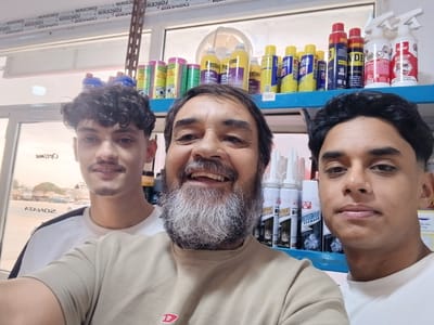 Three people are in a store, likely a auto parts store given the visible products on shelves. Two younger men are on either side of an older man in the center. They are all looking at the camera and smiling. The older man has a prominent grey and white beard. The younger man on the left has curly dark hair and a mustache. The younger man on the right has dark hair and a clean-shaven face. They are all wearing casual clothing.
Behind them, shelves are filled with various automotive products, including cans of oil treatment, fuel treatment, brake cleaner, and other spray cans. Brand names like "Prestone," "Holts," and "W.D.E." are visible. Some of the text on the products is in English, and some appears to be in Arabic. There is also text along the top of the image that reads "FORCEWIN" repeated multiple times.
The scene appears to be indoors with fluorescent lighting, suggesting it is daytime. The overall mood is friendly and casual, with the subjects posing for a selfie. A window in the background shows a glimpse of the outdoors with some buildings and vehicles, and a sign with Arabic text "ATAIO2" which is "OZIA T A" reversed, suggesting it might be a reflection or a reversed sign. Given the mention of Libya, the setting is likely a store in Hun, Libya.