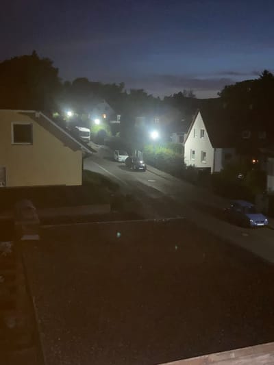 The image depicts a nighttime scene on a street in a residential area of Kassel, Germany. The sky is dark blue, transitioning to lighter hues near the horizon, with some faint clouds visible. Several streetlights illuminate the scene, casting a warm glow on the road and surrounding foliage.
On the left side of the image, a light-yellow house with a dark-framed window is partially visible. Further down the street, a white camper van is parked. Across the street, a small white car is parked near a dense cluster of green bushes. Further up the road, a light blue car is parked on the right side. Several other houses with lit windows are visible in the background, suggesting a populated neighborhood. The street itself is paved and appears to be on a slight incline. The foreground of the image shows a dark, possibly tiled or rooftop surface, with some scattered debris.