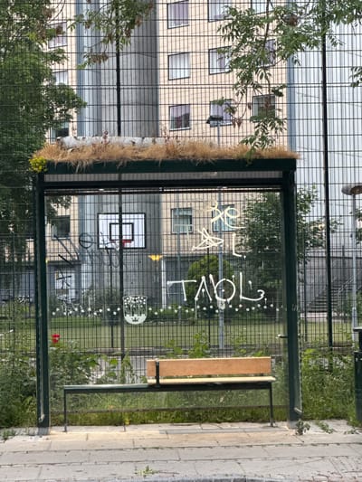 A vertical shot shows a fence surrounding an outdoor sports court. The fence is made of metal mesh and has a dark green frame. A section of the fence is adorned with a decorative element that includes a log wrapped in dry grass and leaves. To the right, a wooden bench is attached to the fence, offering a place to sit.

On the glass panels of the fence, white lettering and graphics are visible. The text appears to read "LIFE IS LOVE." There are also some decorative white dots and a graphic resembling a tree with birds. In the background, through the fence, a basketball hoop and backboard can be seen. Beyond that, a light-colored building with many windows is visible, suggesting an urban or residential setting. Lush green trees and bushes grow around the sports court.

The lighting suggests it is daytime, likely late afternoon or early evening due to the quality of light. The scene appears peaceful and perhaps a bit artistic, given the decorative elements. The overall impression is of an outdoor recreational space with some unique decorative touches. The location is Frederiksberg, Denmark.