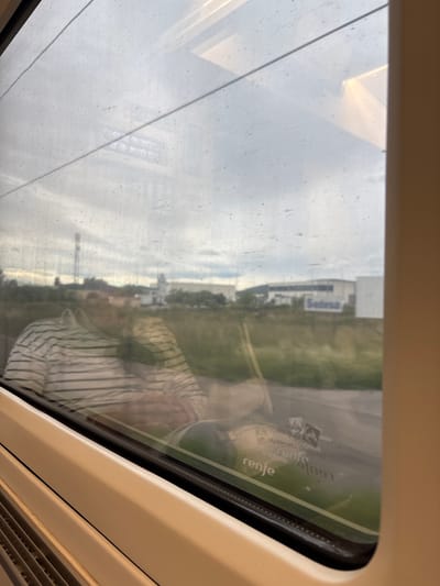 The image shows a view from inside a train, looking through a window. Reflected in the window is a person wearing a striped shirt. The landscape outside the window includes buildings and greenery. A building with "Sedesa" written on it is visible in the distance. A sticker with "renfe" is seen on the window. The location is Alcalà de Xivert, Spain.